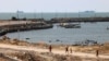 Palestinians walk past a jetty in Gaza City with a view of navy vessels off the coast as part of a humanitarian "maritime corridor" announced by U.S. Central Command on May 17, 2024.
